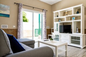 A living room with a sofa, a coffee table, a TV on a white cabinet, and a sliding glass door leading to an outdoor area.