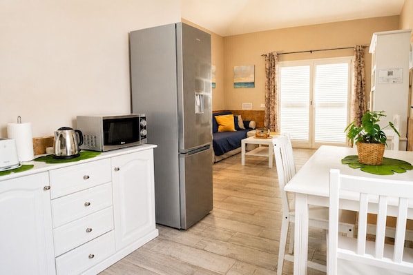 Una cocina compacta con refrigerador, microondas y hervidor, un comedor con mesa y sillas, y una sala de estar con sofá y cortinas.