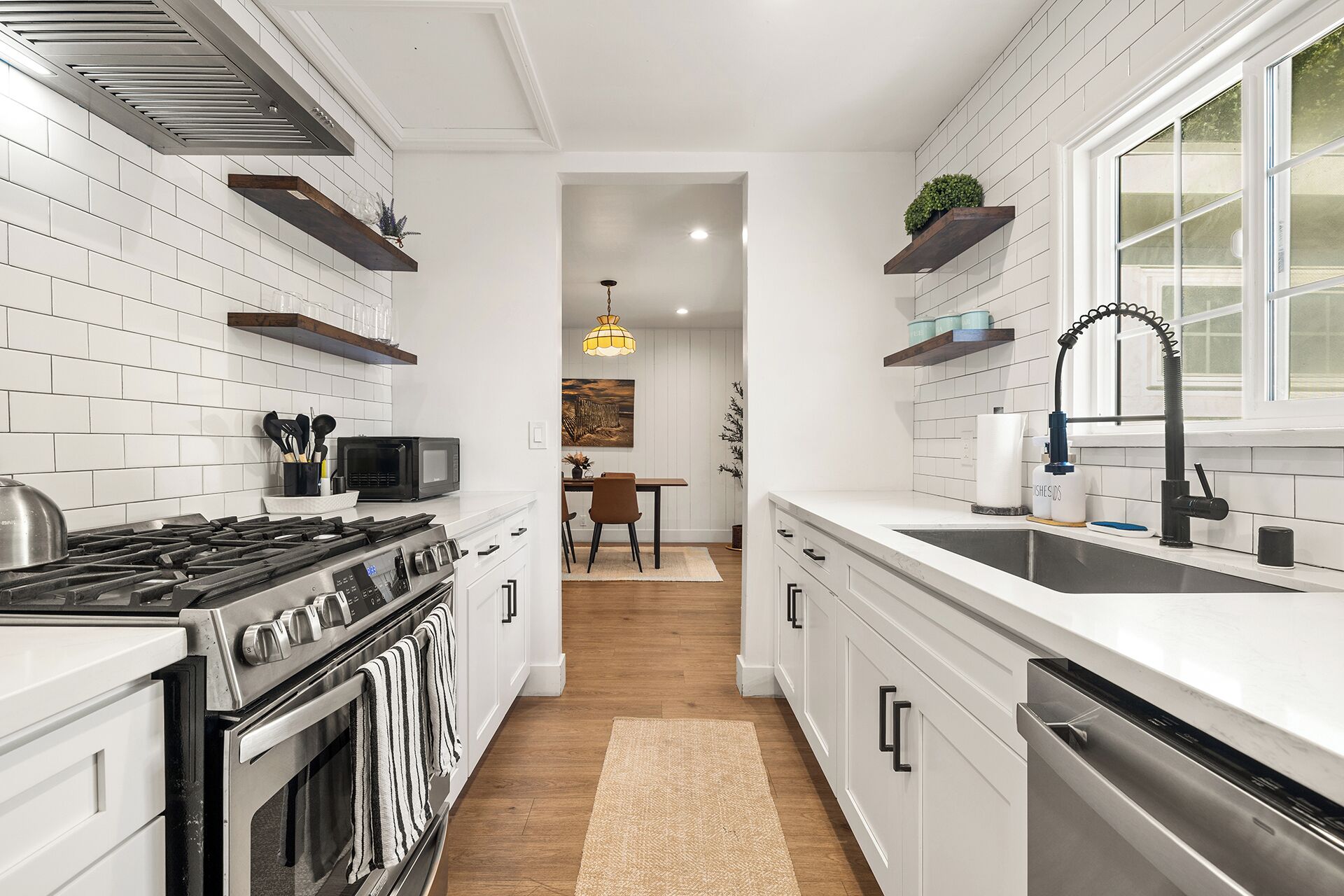 Een moderne keuken met witte kastjes, een gasfornuis, een magnetron en een vaatwasser. Er is een raam met uitzicht op de omgeving, en een eethoek op de achtergrond.