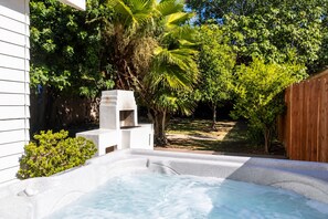 Un bain à remous dans un jardin, avec une clôture en bois, des arbres et un bâtiment blanc.