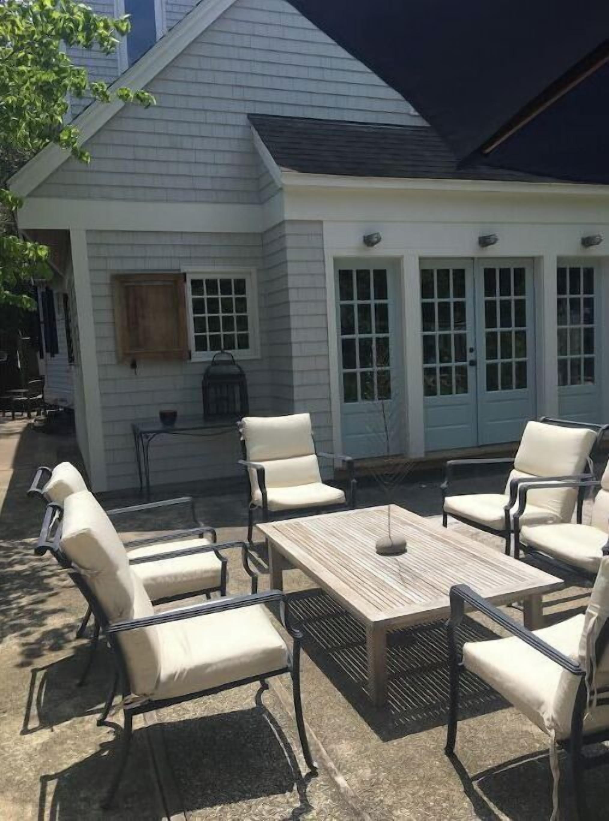 An outdoor patio with a wooden table and six chairs, a small table with a lantern, and a building with light blue doors and windows.