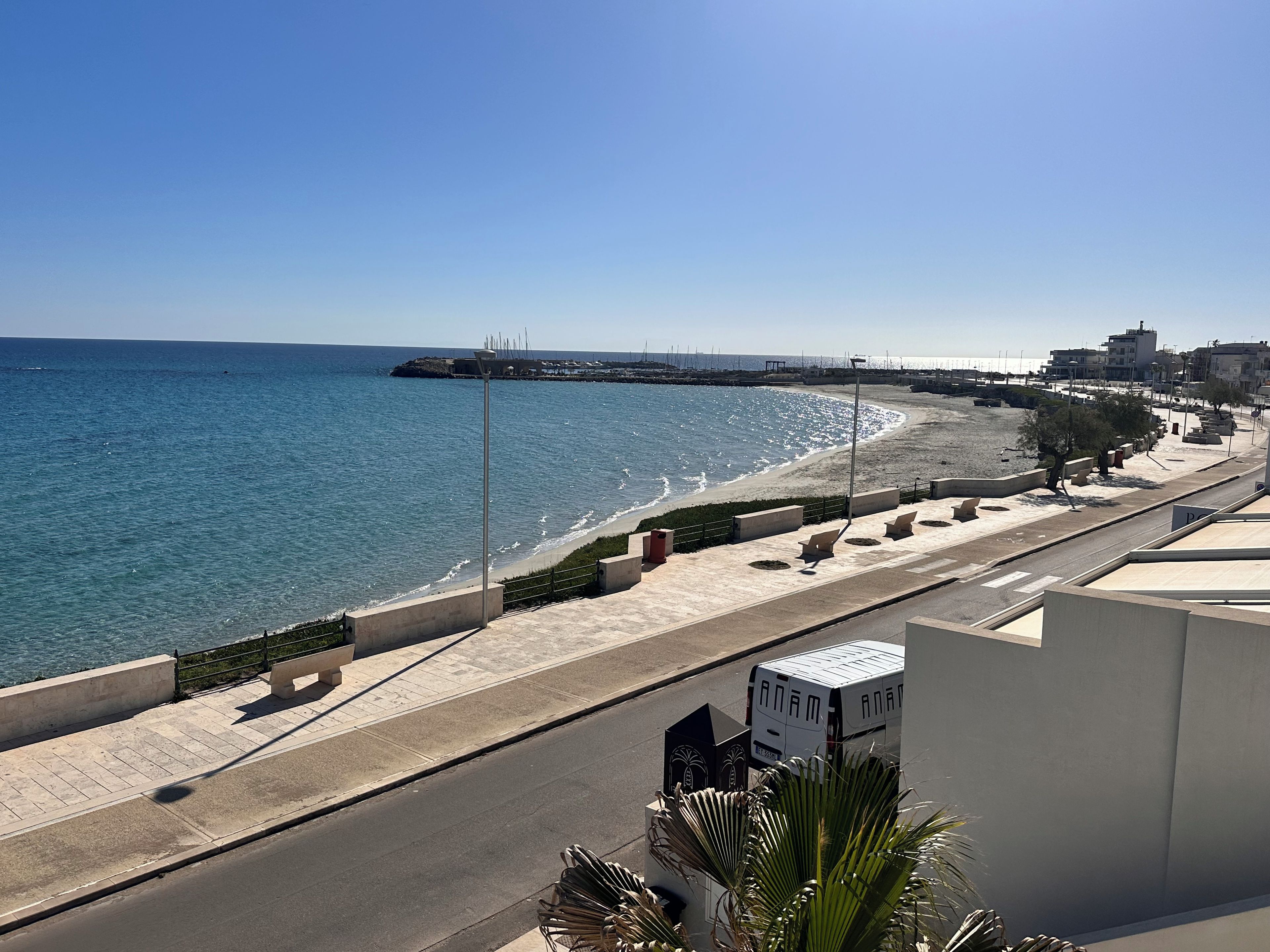 A coastal road with a beach on one side, a clear sky, and buildings in the distance.