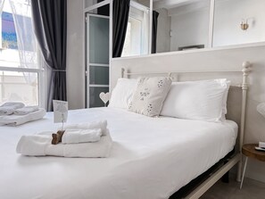 Una cama bien hecha con ropa blanca y almohadas, una mesita de noche con un control remoto y una ventana con cortinas.
