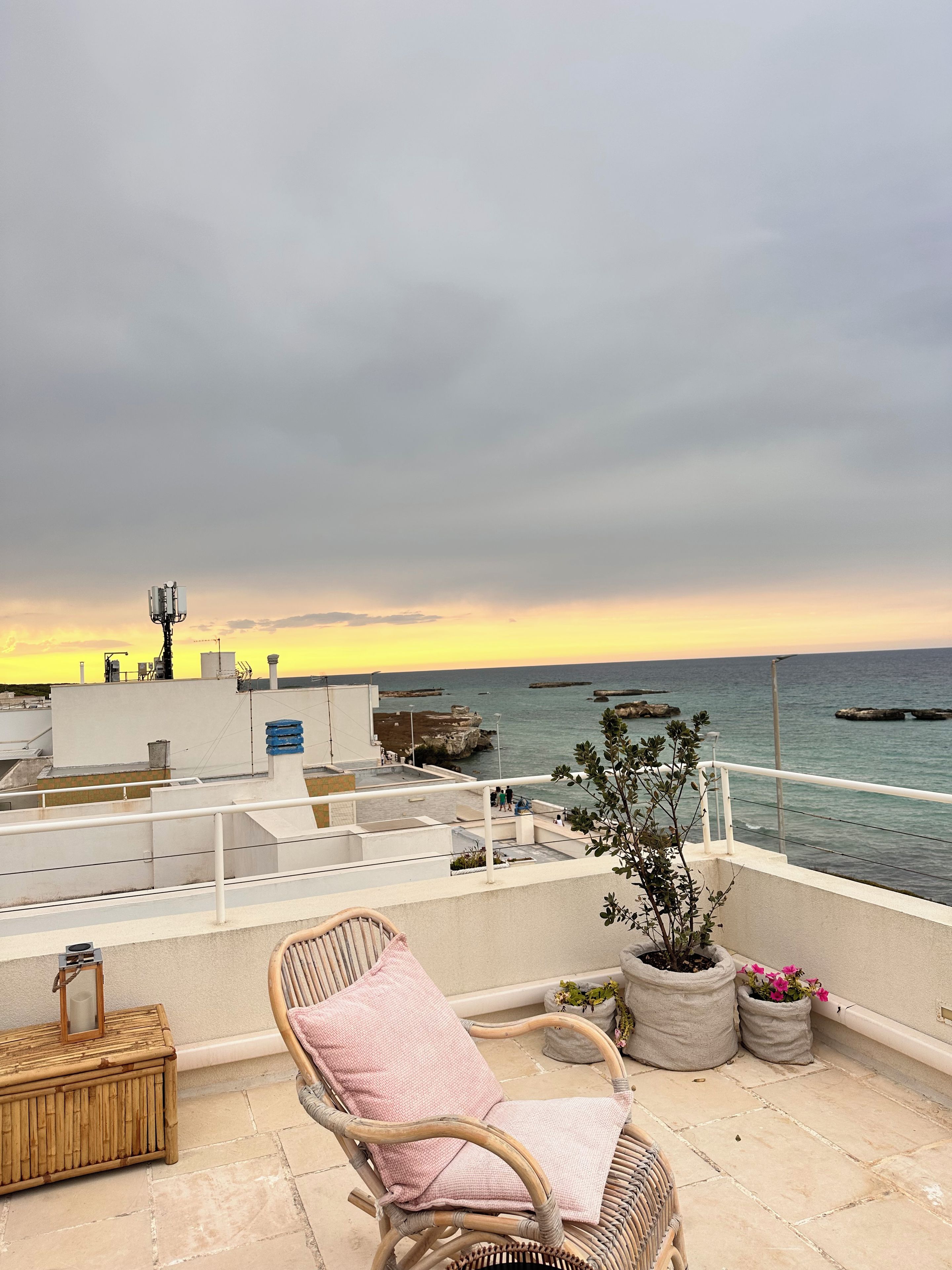 Ein Balkon mit Korbstuhl, Topfpflanzen und Blick auf das Meer bei Sonnenuntergang.