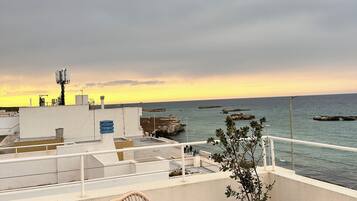 Ein Balkon mit Korbstuhl, Topfpflanzen und Blick auf das Meer bei Sonnenuntergang.