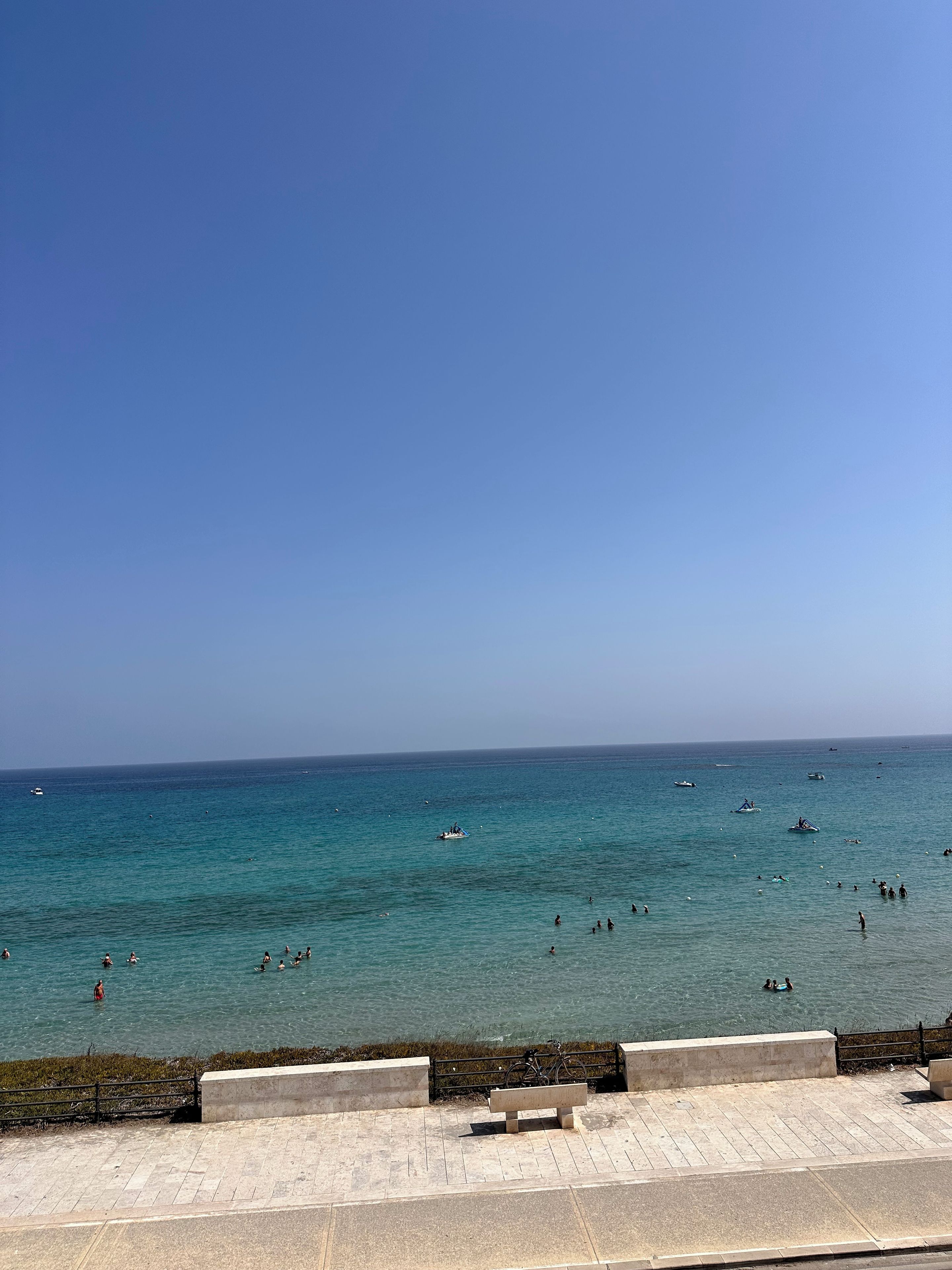 Een uitzicht op de kust, met blauwe lucht, rustige zee en een promenade met een bankje.
