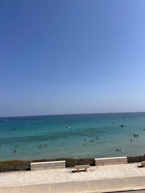 A coastal view with a clear sky, calm sea, and a promenade with a bench.