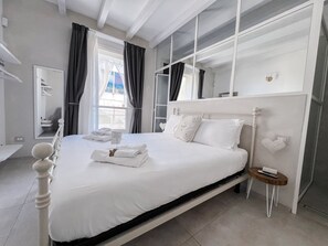 A neatly made bed with white linens and pillows, a bedside table with a remote control, and a window with curtains.