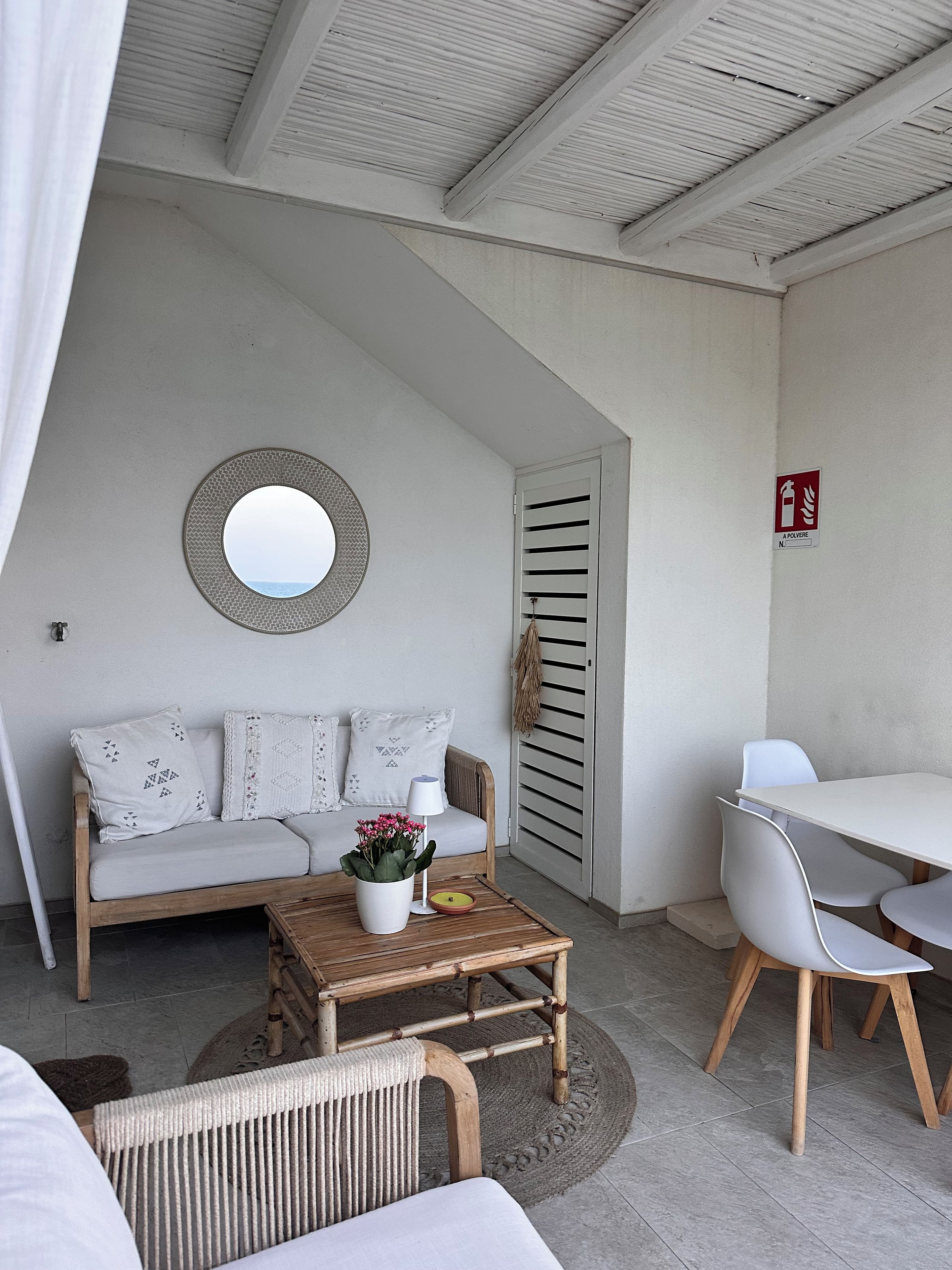 Quarto com sofá branco, espelho redondo, mesa de madeira com planta em vaso e cadeira branca.