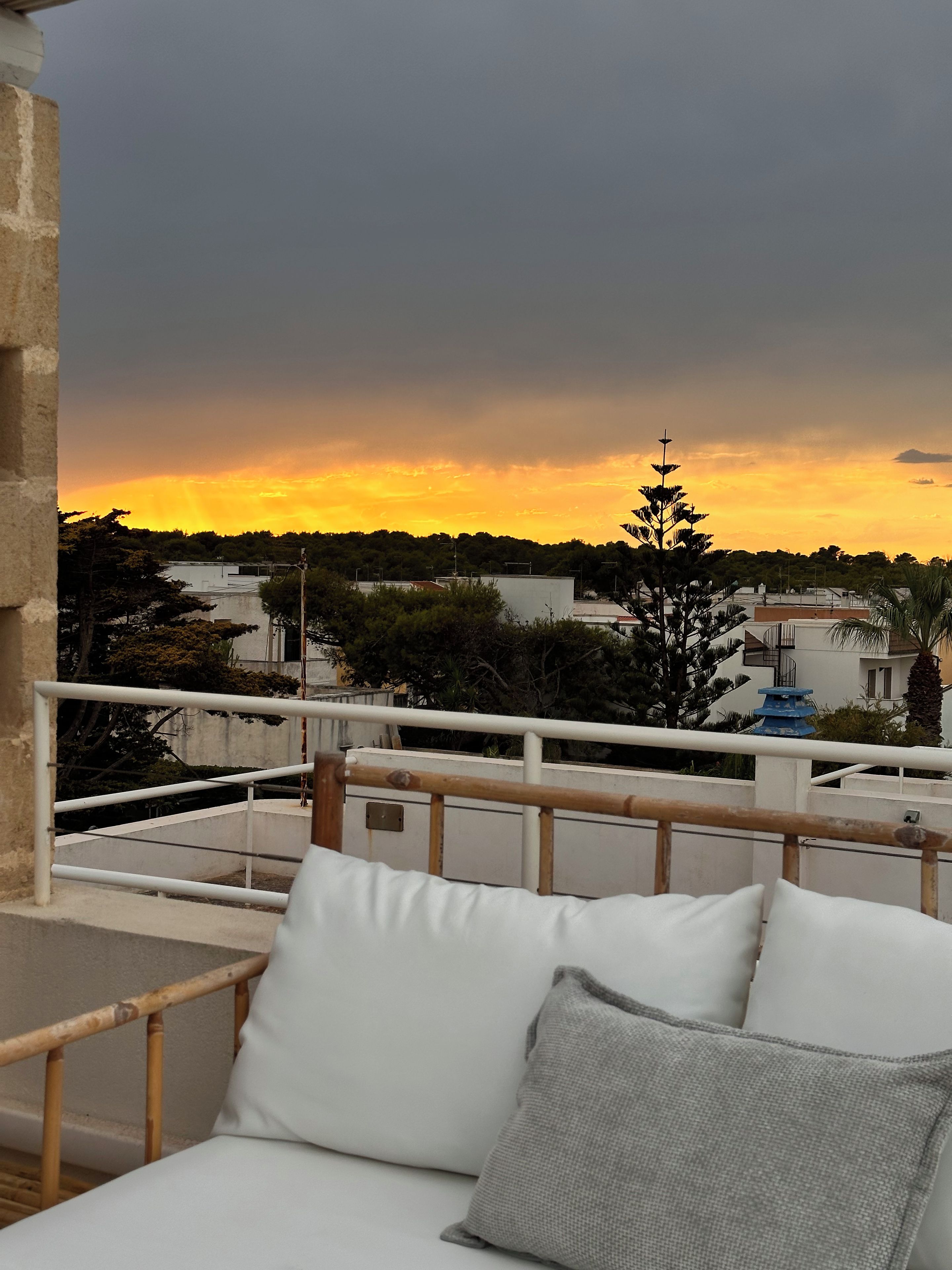 Ein Balkon mit einer weißen Sitzgelegenheit, einem Geländer und Blick auf Gebäude und Bäume unter einem Sonnenuntergangshimmel.
