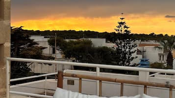 Ein Balkon mit einer weißen Sitzgelegenheit, einem Geländer und Blick auf Gebäude und Bäume unter einem Sonnenuntergangshimmel.