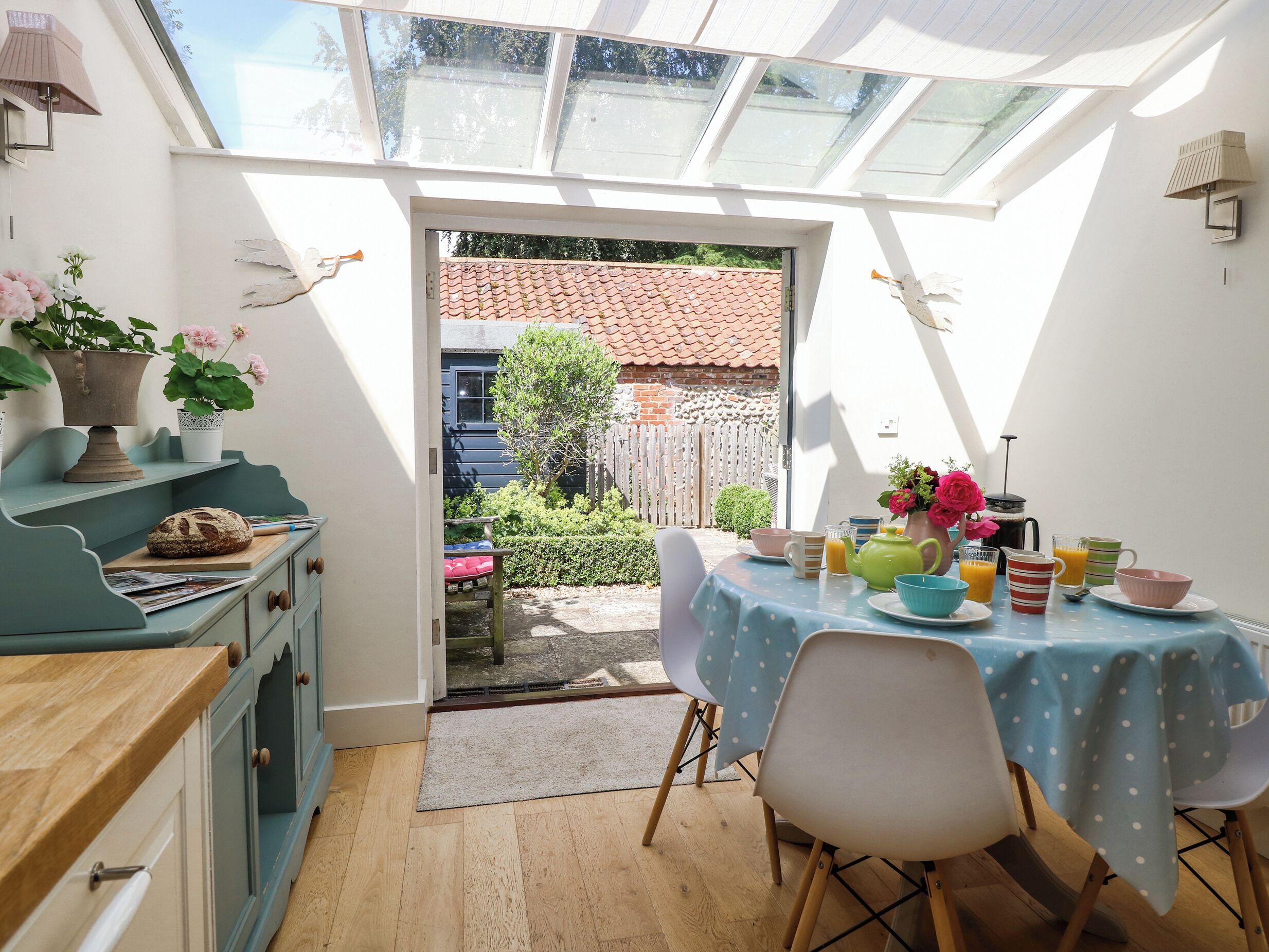 Un coin petit-déjeuner accueillant avec une table dressée pour deux, un petit espace cuisine avec un pain et une plante, et une vue sur un jardin par une porte vitrée.