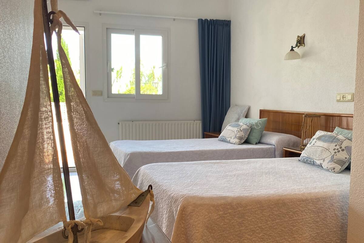 Ein Hotelzimmer mit zwei Betten, einem Holztisch, einem dekorativen Segel, einem fenster mit Vorhängen und einer Wandleuchte.