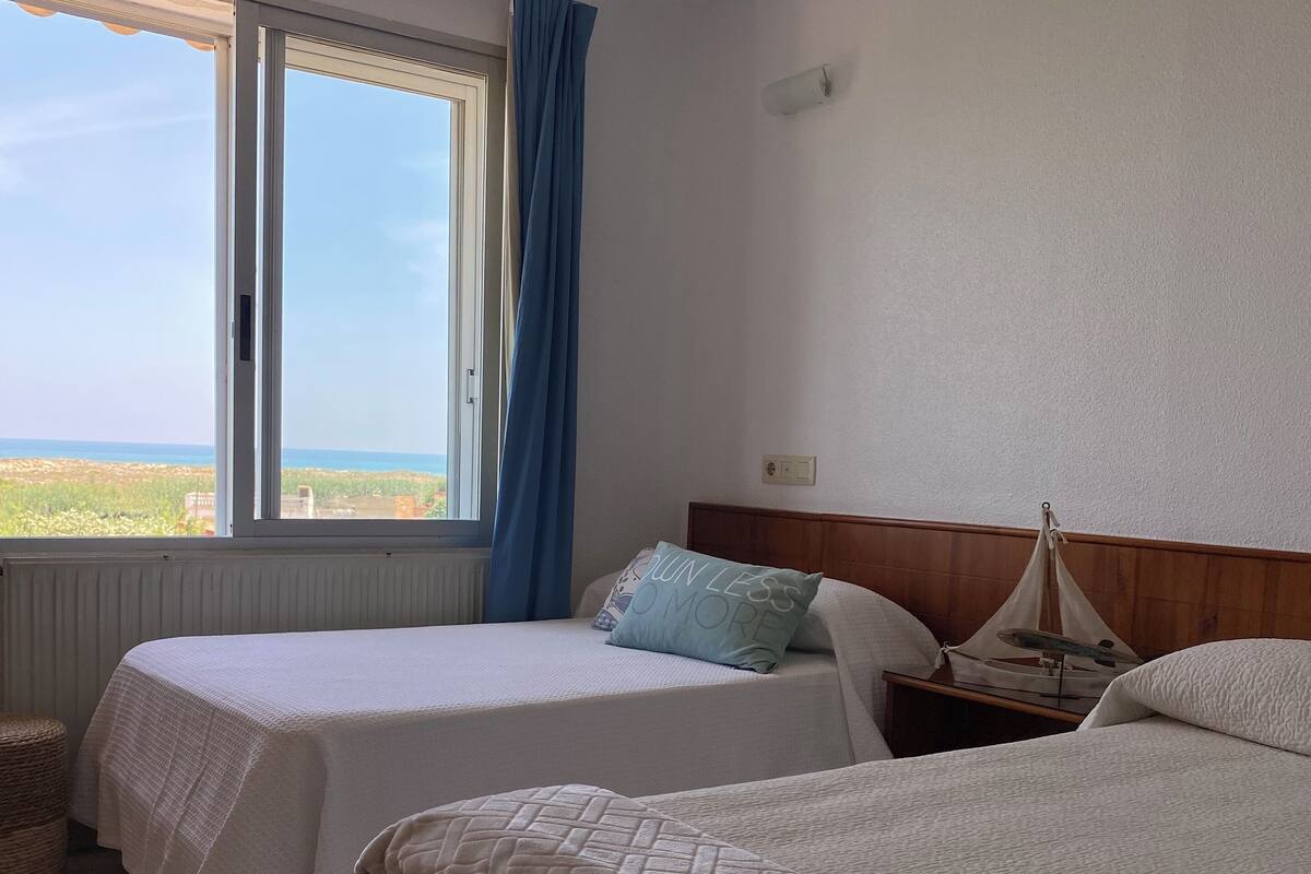 Ein Hotelzimmer mit zwei Betten, einem Fenster mit Meerblick, einem hölzernen Kopfteil und einem kleinen Nachttisch mit Lampe.