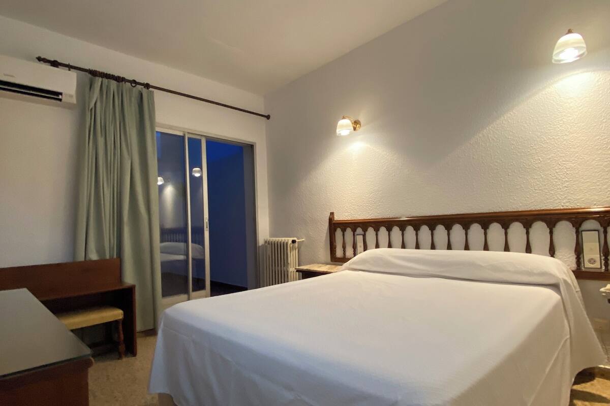 Ein Hotelzimmer mit Bett, Schreibtisch, Sitzbank, Heizkörper und einem fenster mit Vorhängen.