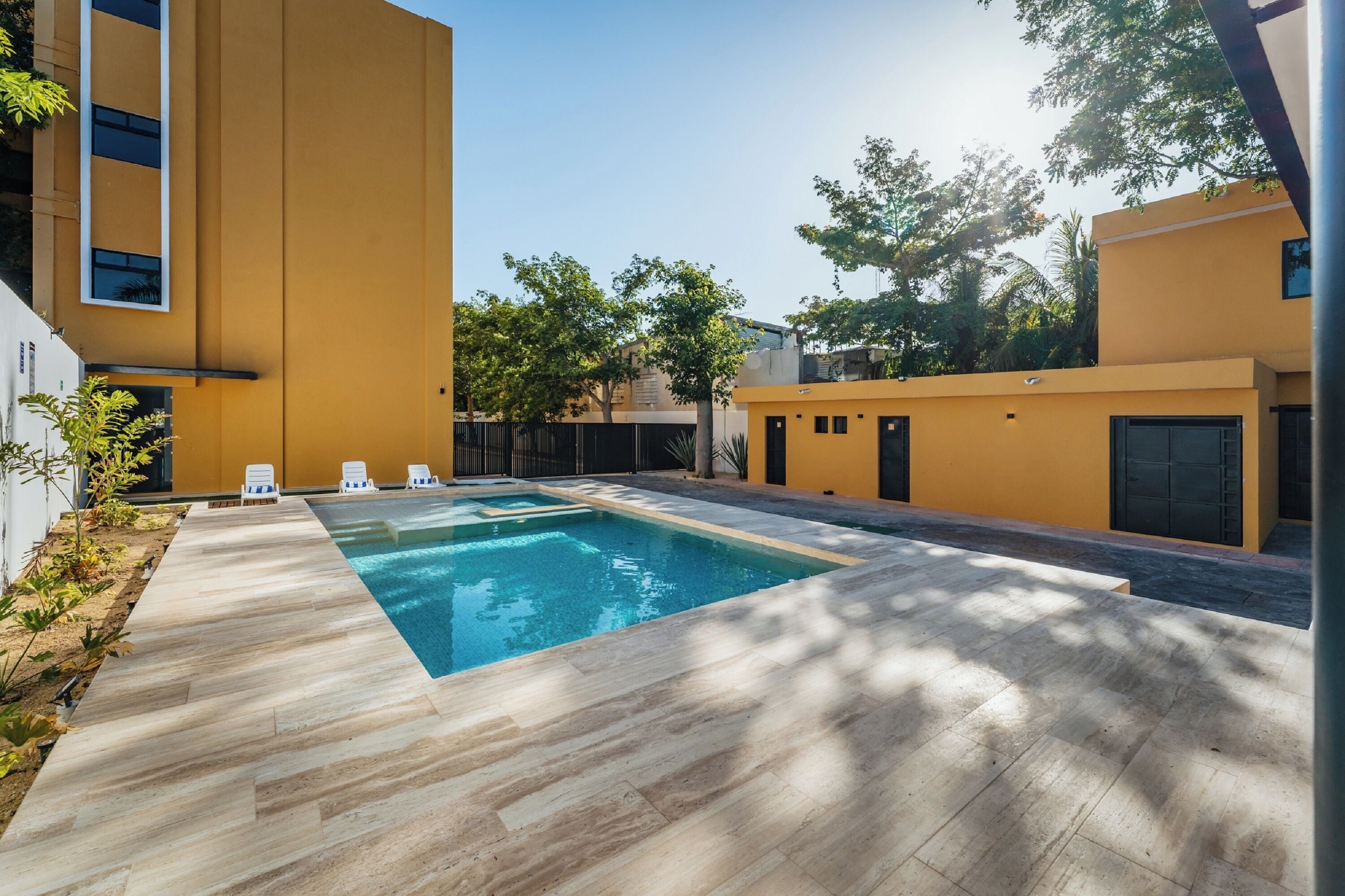 Un área de piscina exterior moderna con una terraza de madera, sillas de descanso y un edificio amarillo al fondo.