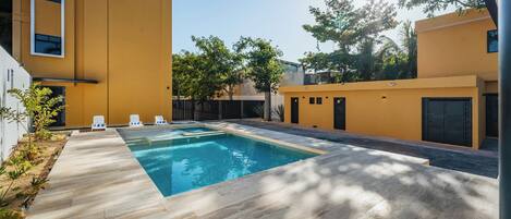 Un área de piscina exterior moderna con una terraza de madera, sillas de descanso y un edificio amarillo al fondo.