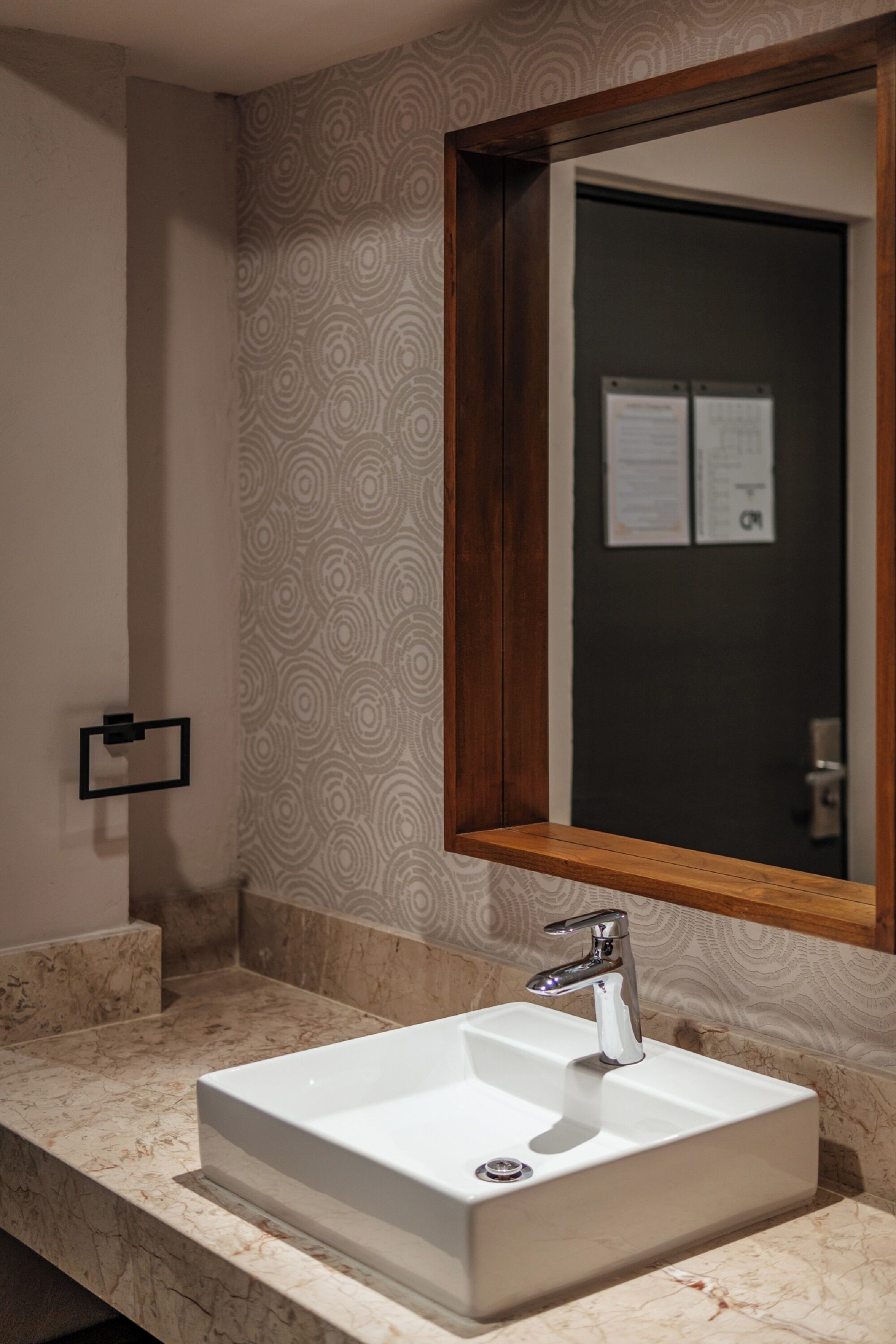 A bathroom with a rectangular sink, a wooden-framed mirror, and patterned wallpaper.
