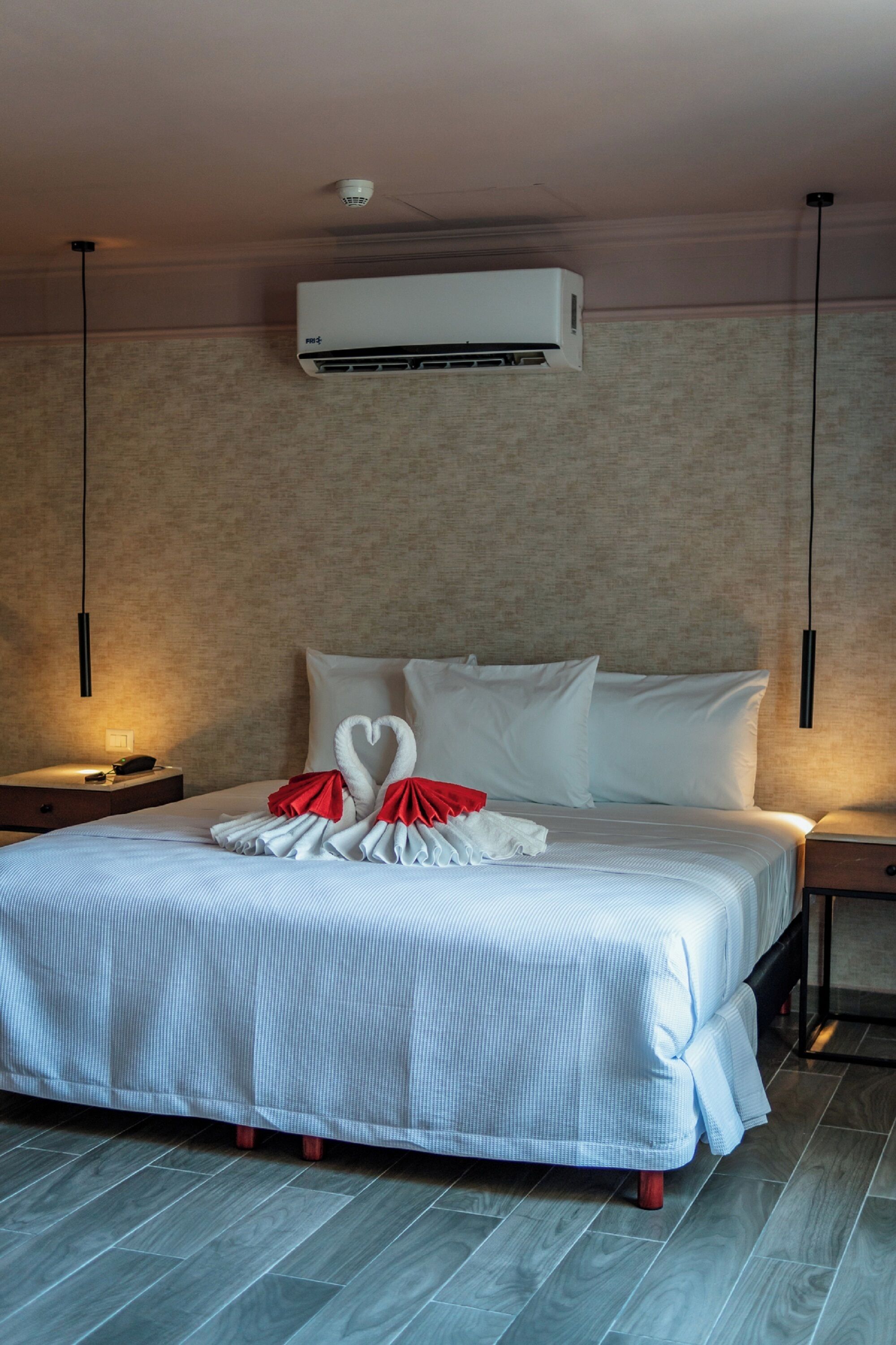 A neatly made bed with white linens and red accents, a wall-mounted air conditioner, and a textured wall.