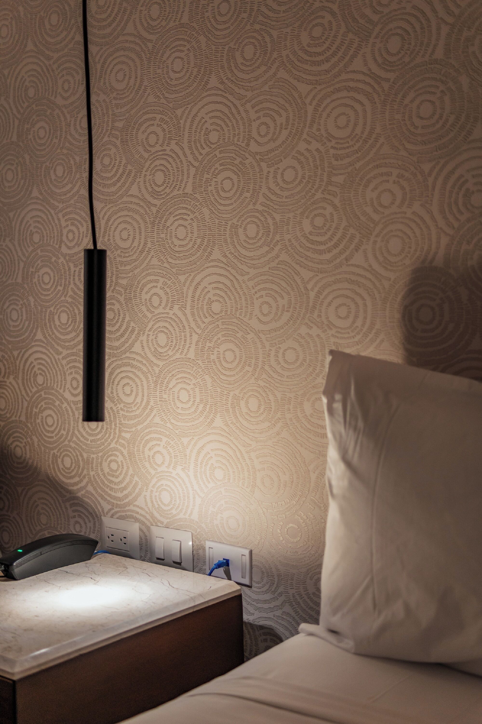 Une chambre d’hôtel avec un papier peint à motifs, une table de chevet avec une lampe et un oreiller blanc.
