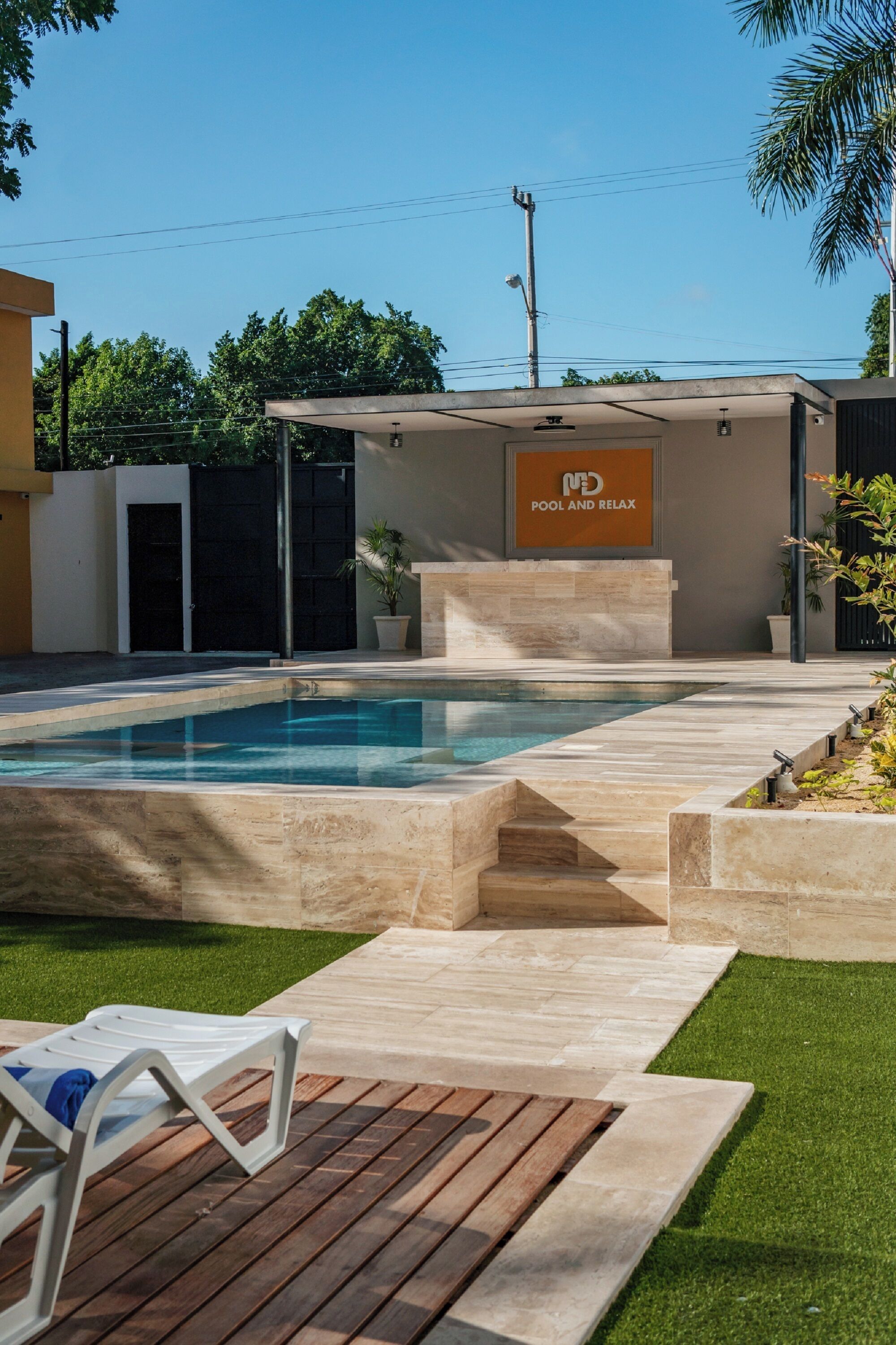 A modern outdoor pool area with a clear blue pool, a lounge chair, and a sign reading "POOL AND RELAX".
