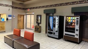 A lobby with a reception desk, a seating area with two cushioned chairs, a refreshment vending machine, and a snack vending machine. - Studio 6 Suites West Memphis, AR - Memphis (West Memphis)