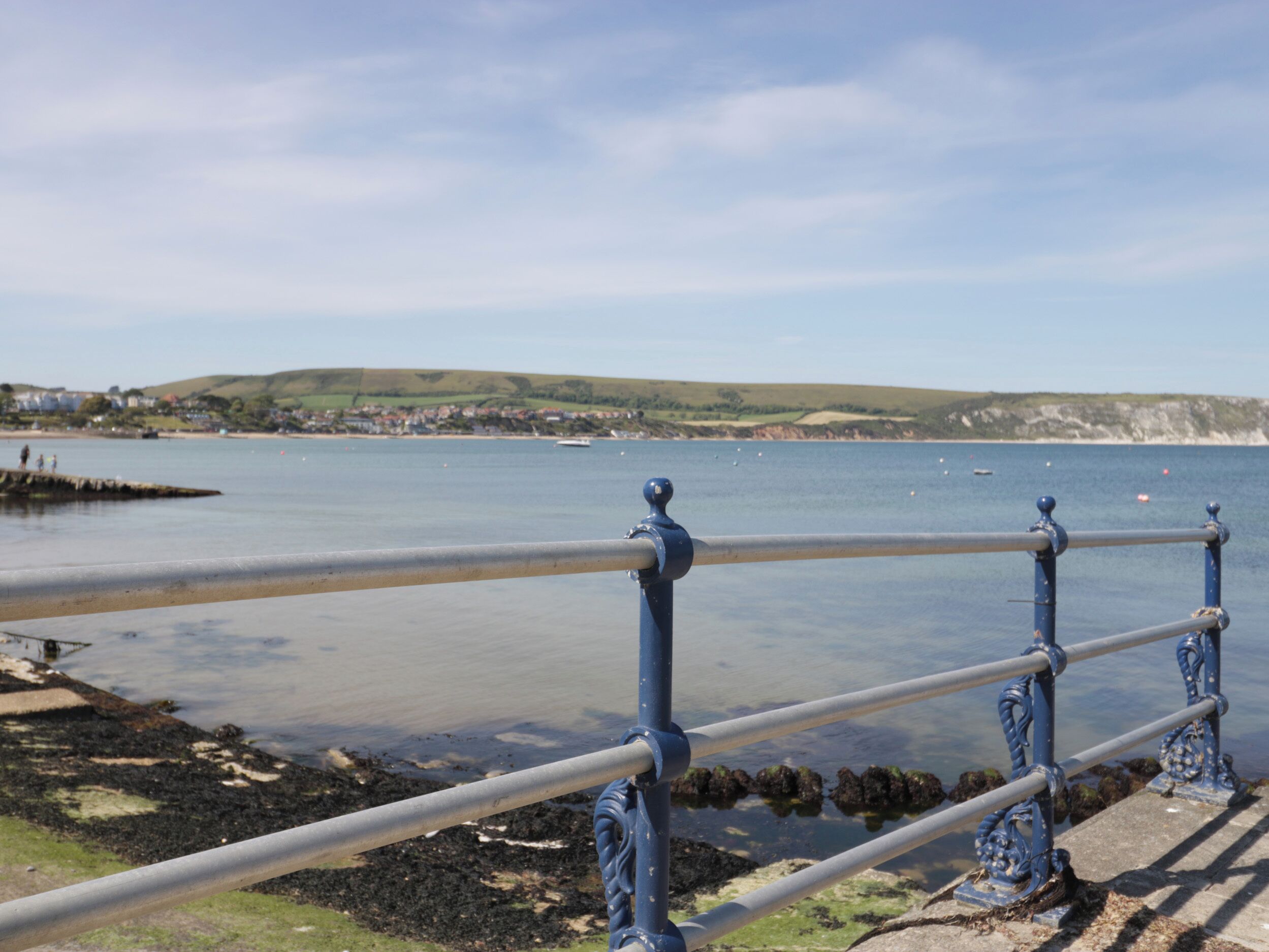 A coastal view with a metal railing, calm water, and a distant shoreline with buildings.