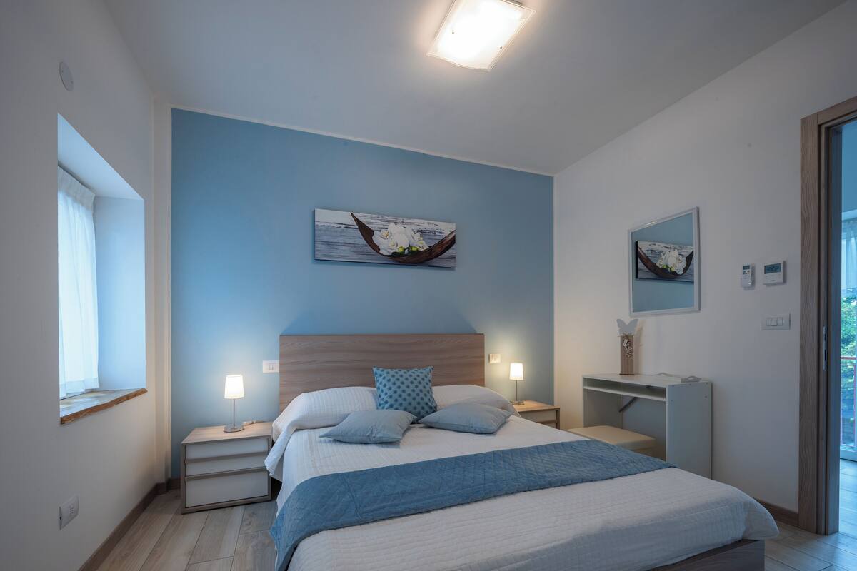 A modern bedroom with a large bed, bedside tables, a nightstand, a desk, and a window.