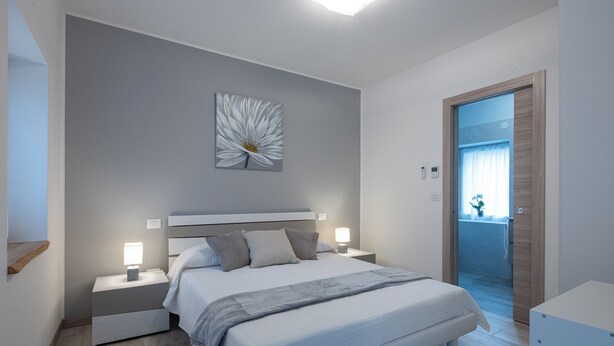 A modern bedroom with a large bed, bedside tables with lamps, a wall-mounted light fixture, and a door leading to another room. - Vigna degli Angeli (Ragogna)