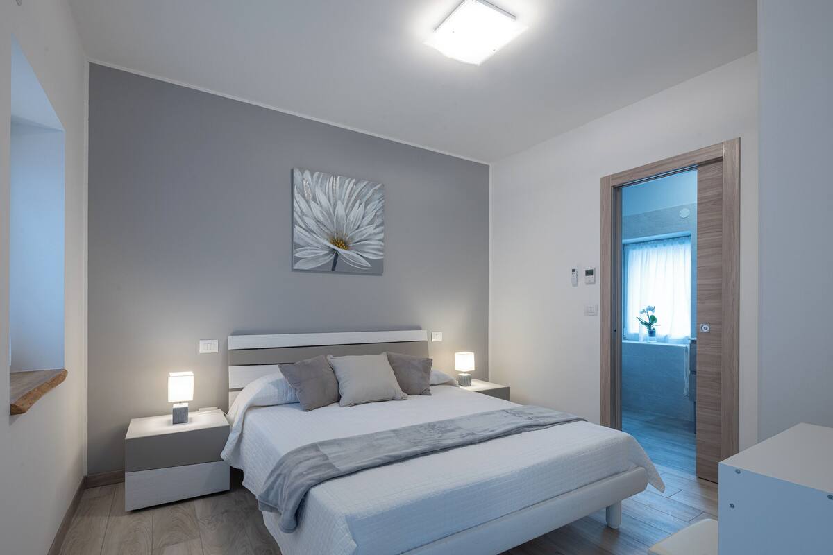 A modern bedroom with a large bed, bedside tables with lamps, a wall-mounted light fixture, and a door leading to another room.