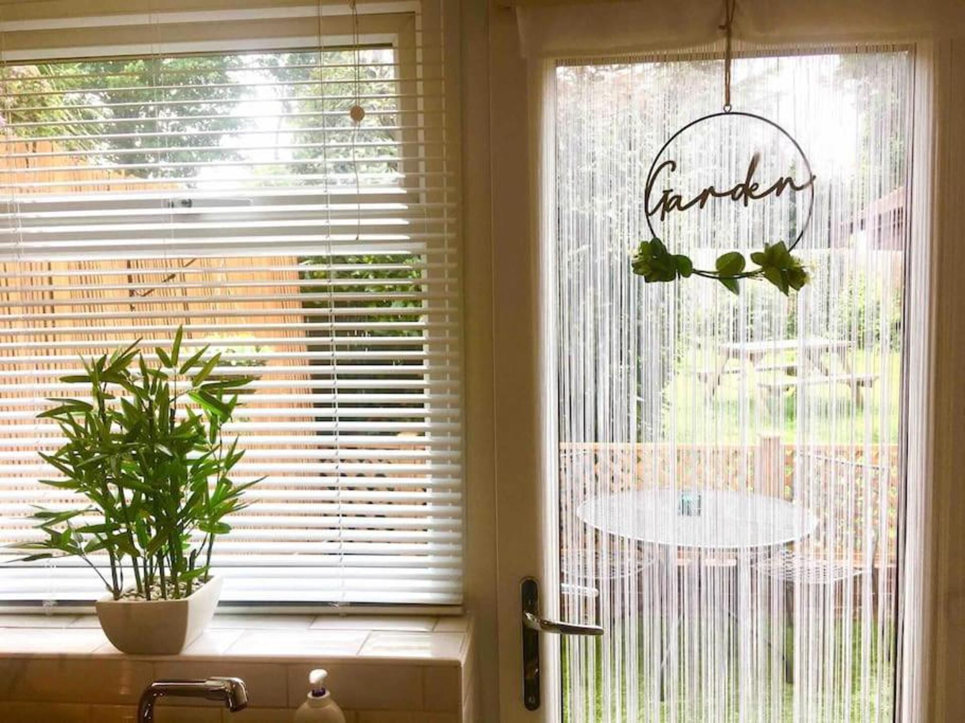 Ein Zimmer mit Fenster, ausgestattet mit weißen Jalousien, einem Blumentopf auf dem Fensterbrett und einer Tür mit einem runden Schild, auf dem "Garten" steht.