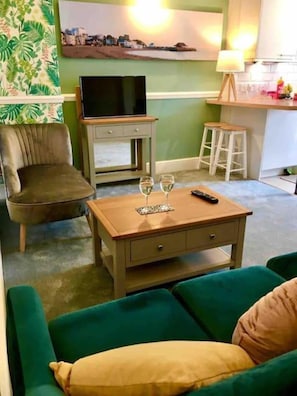 Un salon avec un canapé vert, un fauteuil marron, une table basse en bois sur laquelle se trouvent une télécommande et deux verres, un téléviseur posé sur un meuble et un tableau accroché au mur.