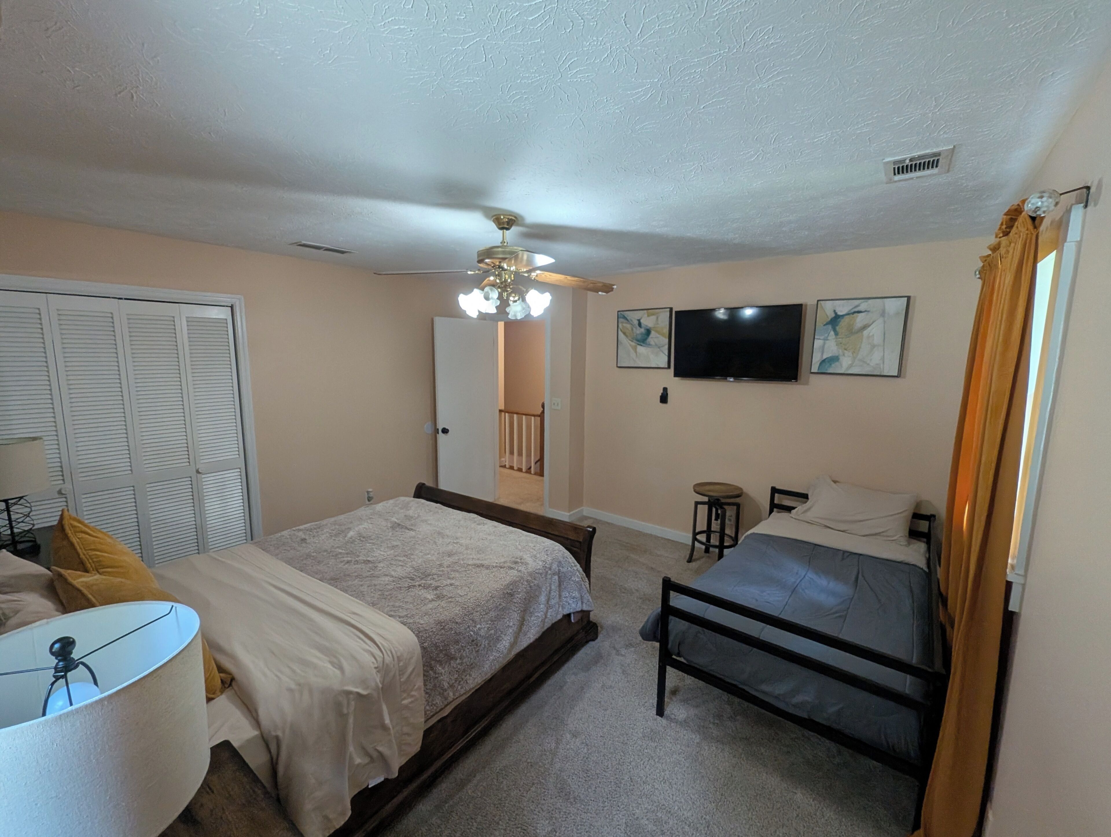 Ein Schlafzimmer mit einem Bett, einem Schreibtisch mit Lampe, einem Deckenventilator, einem Fernseher und Kunstwerken an der Wand.