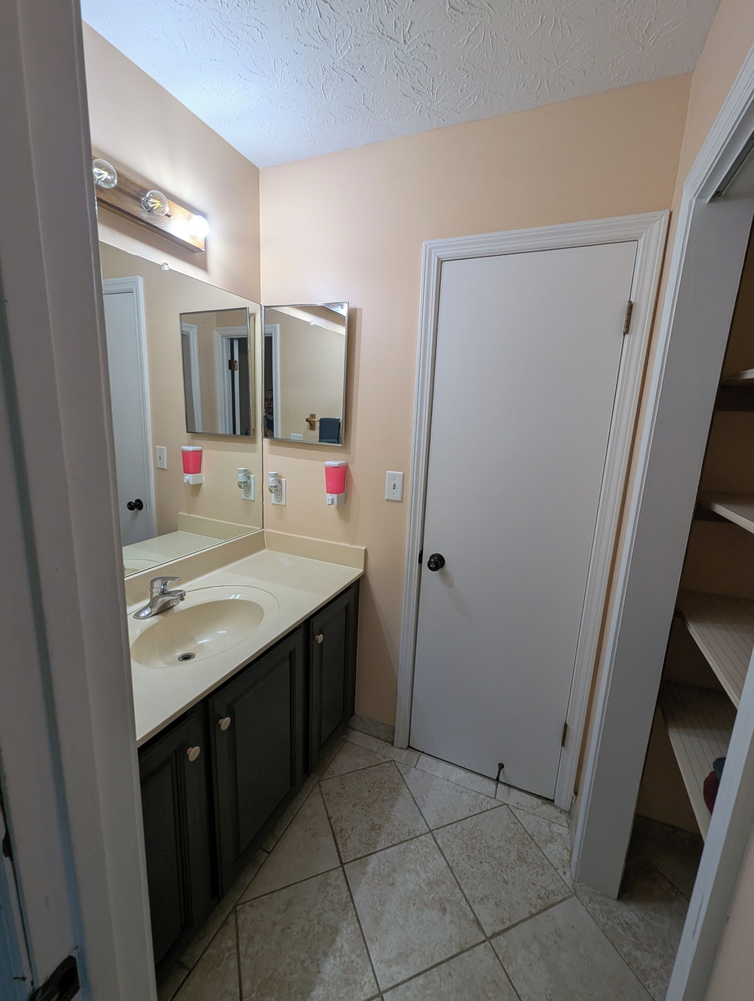 Un baño con lavamanos, dos espejos, una puerta y piso de cerámica.