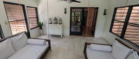 Une pièce spacieuse avec deux canapés blancs, un ventilateur de plafond, une commode blanche et une vue sur l’extérieur à travers une porte vitrée.