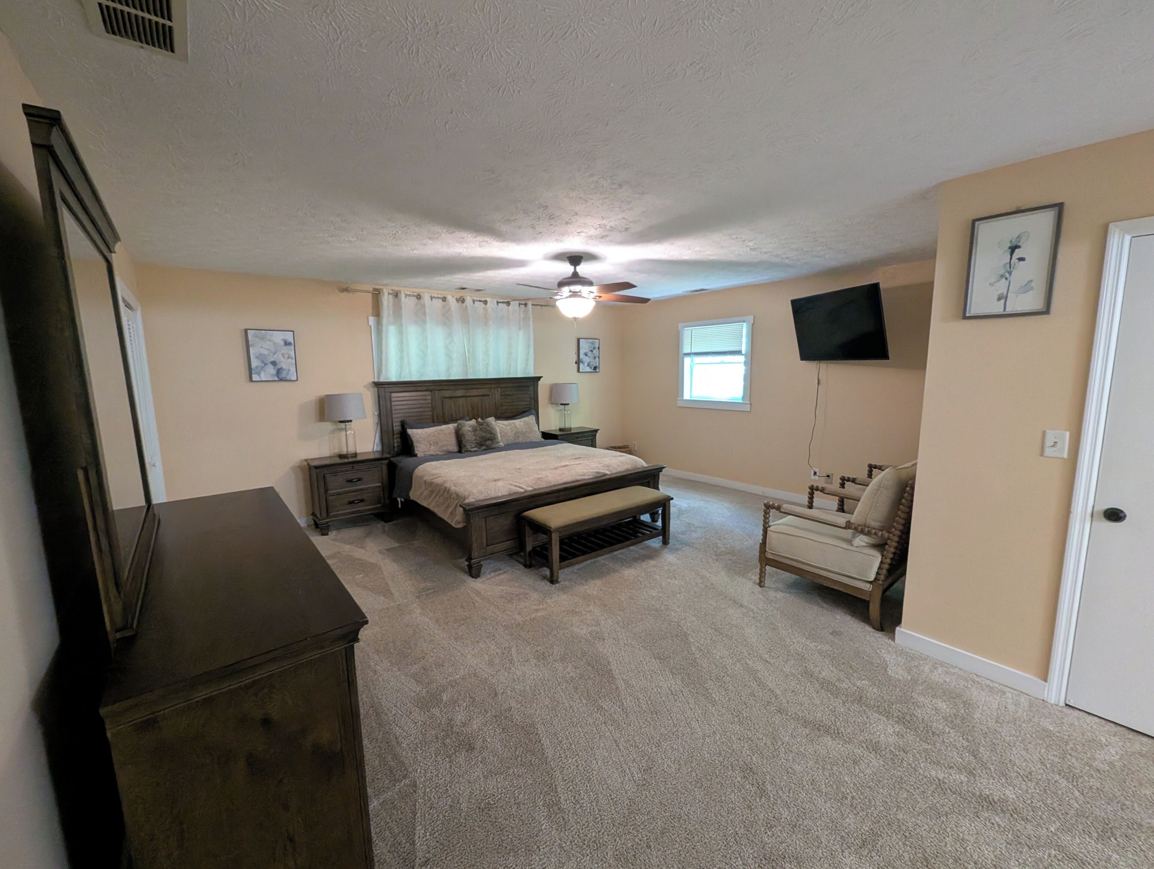 A bedroom with a bed, bedside tables, a chair, a dresser, a ceiling fan, and a TV mounted on the wall.