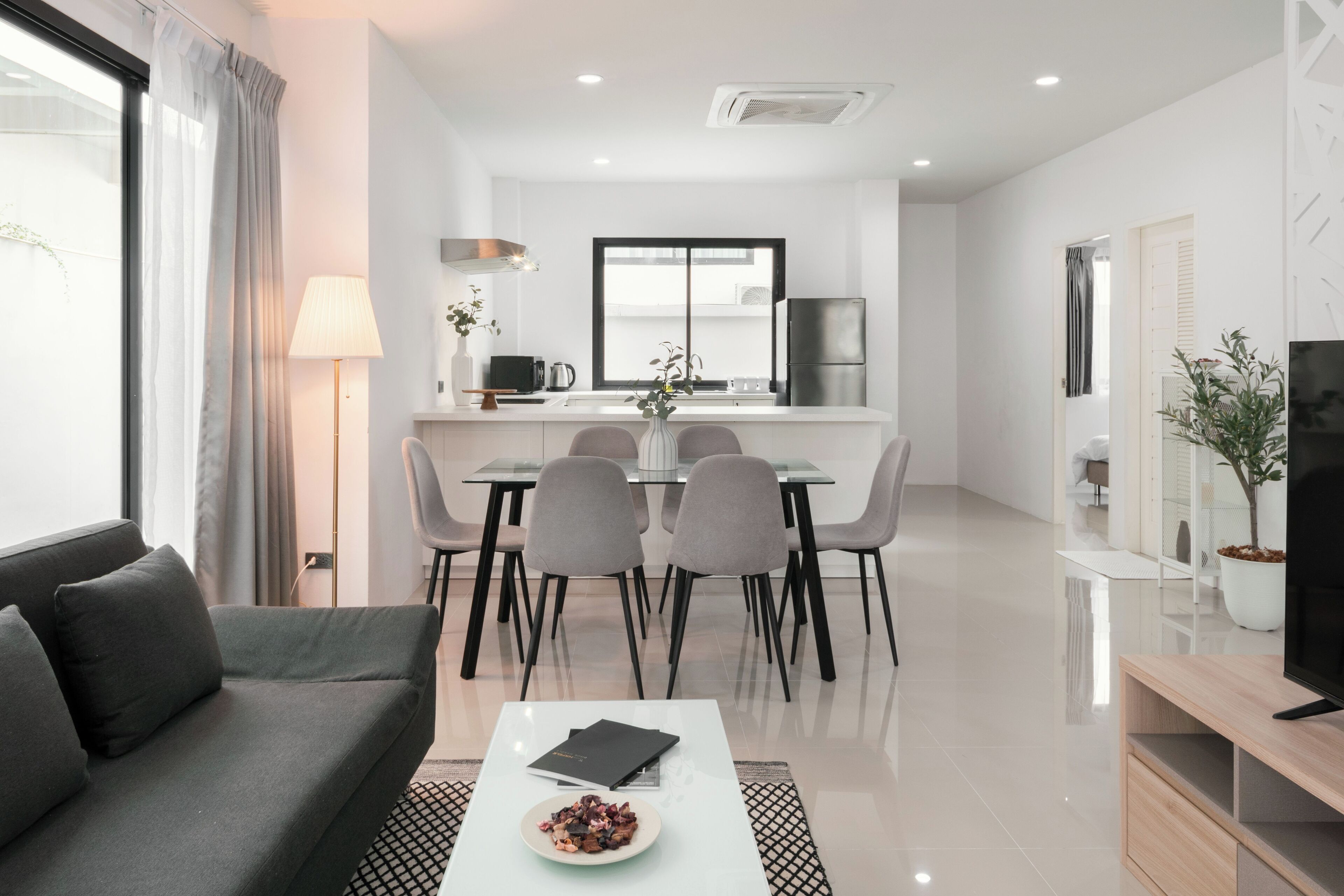 A modern living room with a sofa, a coffee table with a plate of food, a dining area with a table and chairs, a kitchen with a refrigerator, and a TV on a stand.