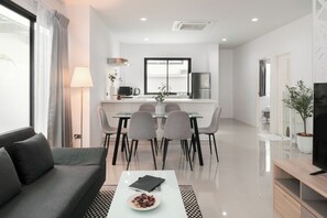 A modern living room with a sofa, a coffee table with a plate of food, a dining area with a table and chairs, a kitchen with a refrigerator, and a TV on a stand. - Chao Wilai Residence | Naiyang Beach | HKT Airport (Sa Khu)
