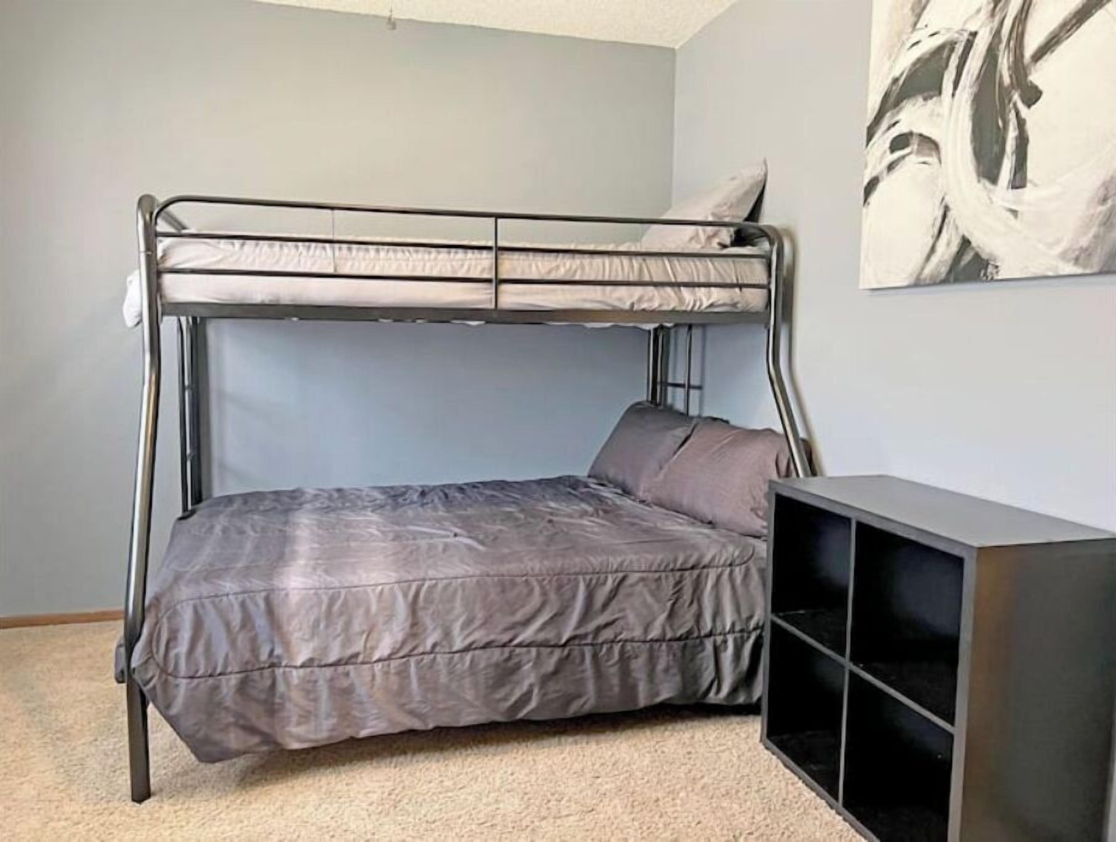 A bunk bed with a grey mattress and pillows, a black TV stand, and a painting on the wall.