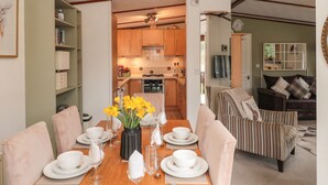 Un coin repas avec une table en bois dressée pour le repas, entourée de chaises rembourrées, un vase avec des fleurs jaunes et une cuisine en arrière-plan.