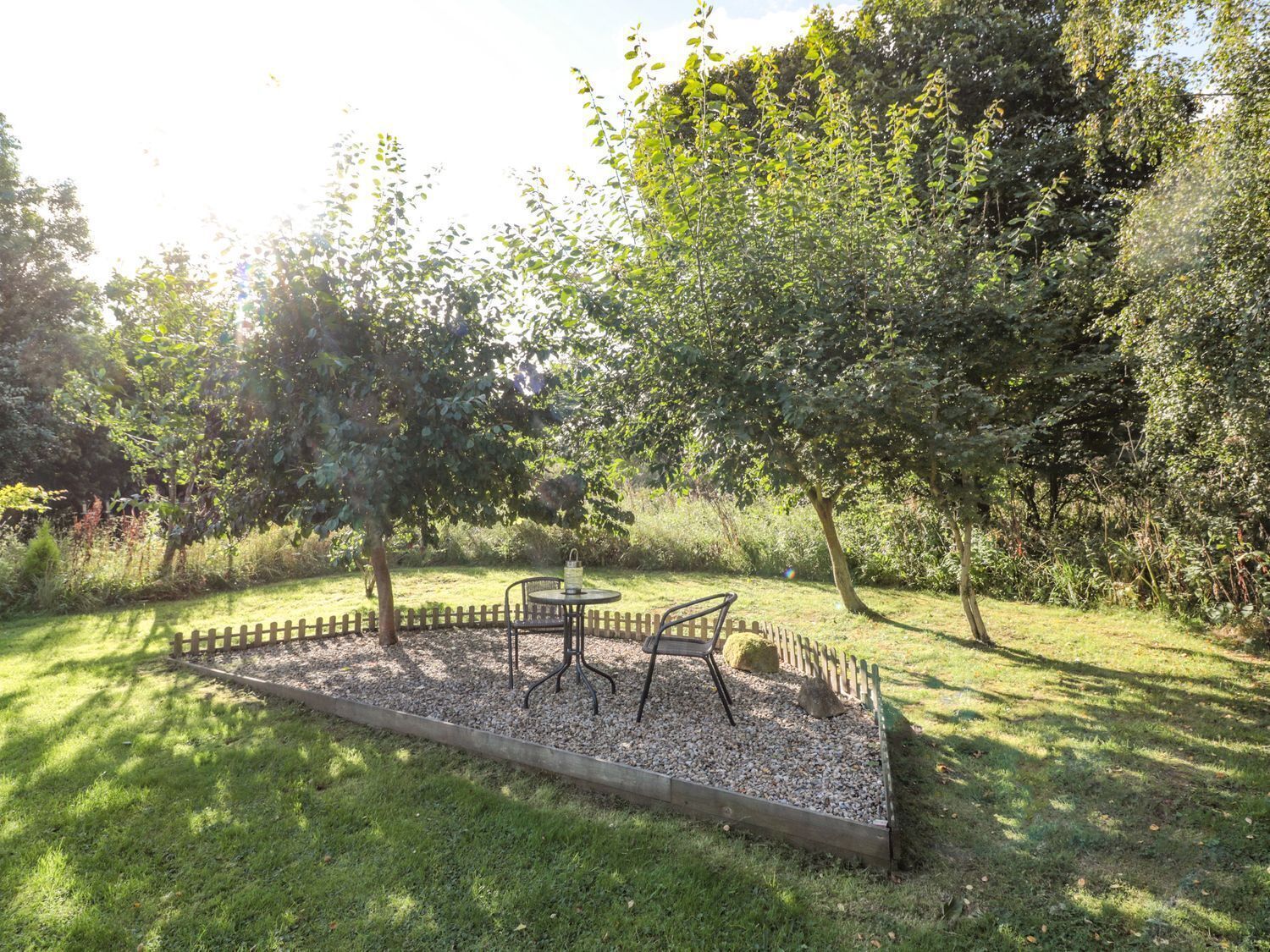 Een tuin met een metalen tafel en stoelen, omgeven door een grindpad, met bomen en een houten omheining op de achtergrond.