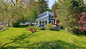 A modern two-story house with a well-maintained lawn and garden, surrounded by trees and shrubs. - Cozy Cottage by Lake Simcoe | Private Dock & Kayaks Included (Brechin)