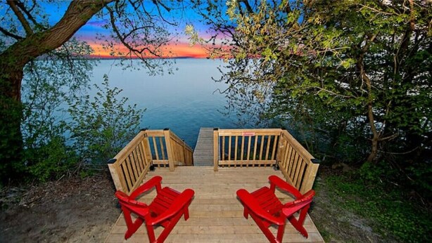 A wooden deck with two red Adirondack chairs facing a calm lake at sunset. - Cozy Cottage by Lake Simcoe | Private Dock & Kayaks Included (Brechin)