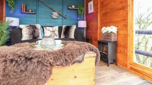 Interior acogedor de una cabaña con techo de madera, pared azul decorativa, sofá con cojines estampados, manta de piel, mesita con un cuenco decorativo y ventana con vistas a árboles.