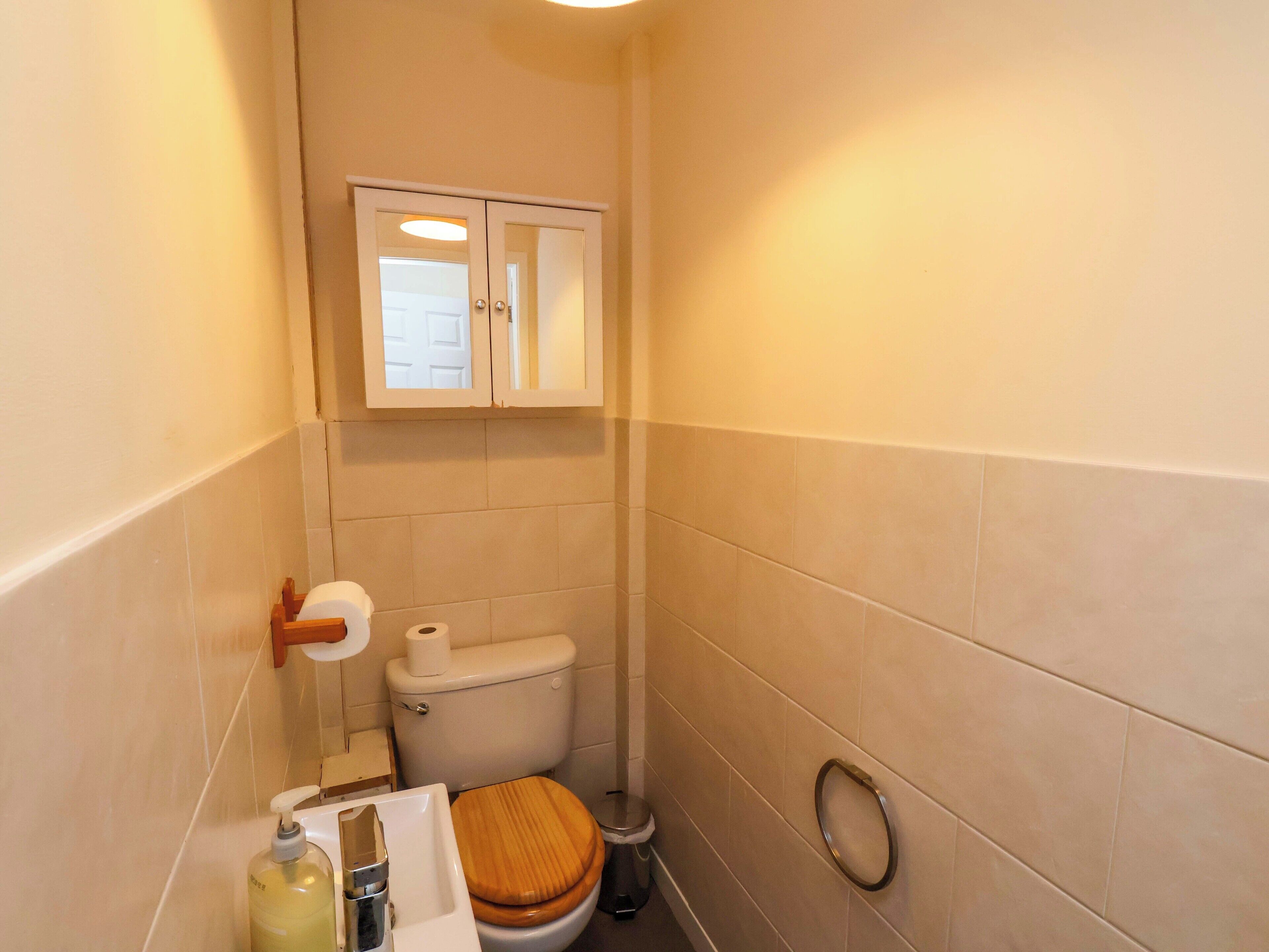Un baño pequeño con inodoro, un asiento de madera, un secador de manos, dispensador de jabón y un armario sobre el inodoro.