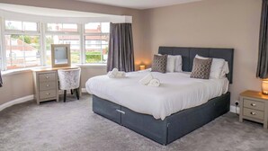 A bedroom with a large bed, a desk, and a chair. There is a window with curtains, a bedside table with a lamp, and a ceiling light. - Hazelgarth House (Filey)