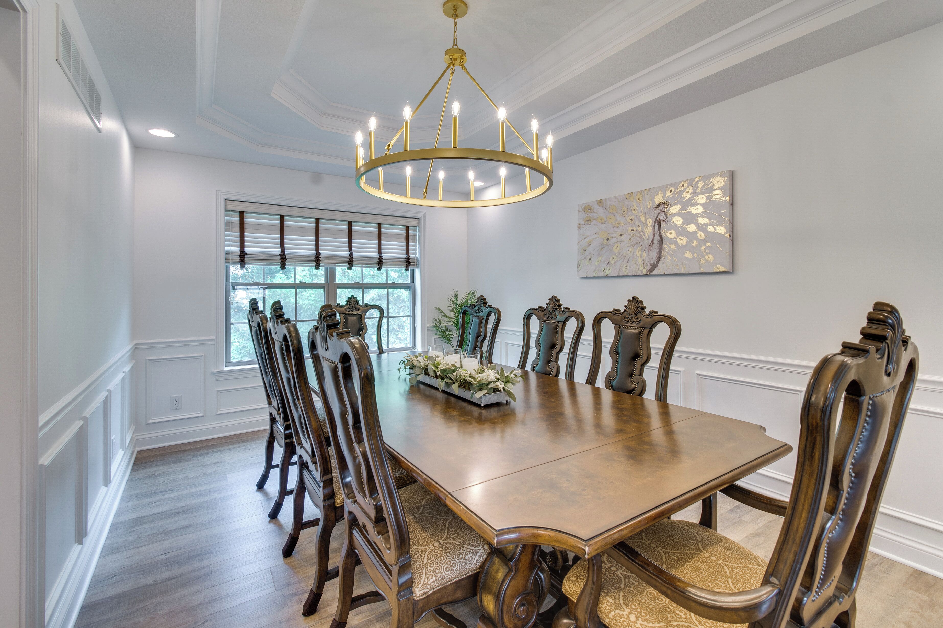 Une salle à manger avec une grande table en bois, des chaises décorées, un lustre et une fenêtre avec des stores.