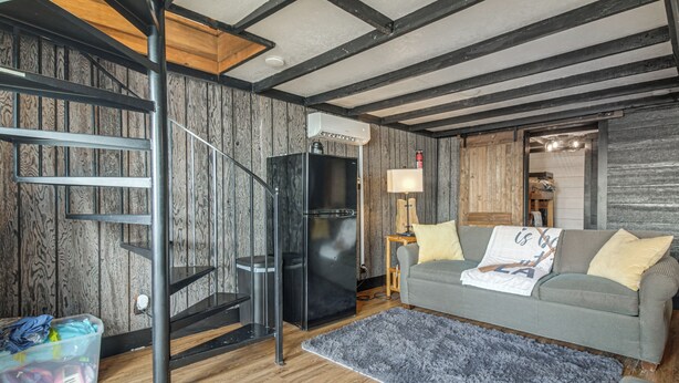 Ein modernes Wohnzimmer mit Holzboden, einem grauen Sofa, einem schwarzen Kühlschrank, einer Holztreppe und einem kleinen Tisch mit Lampe.