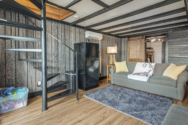 A modern living room with a wooden floor, a grey sofa, a black refrigerator, a wooden staircase, and a small table with a lamp. - Private Hot Tub & Dock: Lakefront Grove Retreat! (Grove)