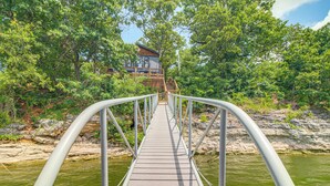 Eine Holzbrücke mit Metallgeländern führt zu einem Gebäude, das von Bäumen und einem Gewässer umgeben ist.
